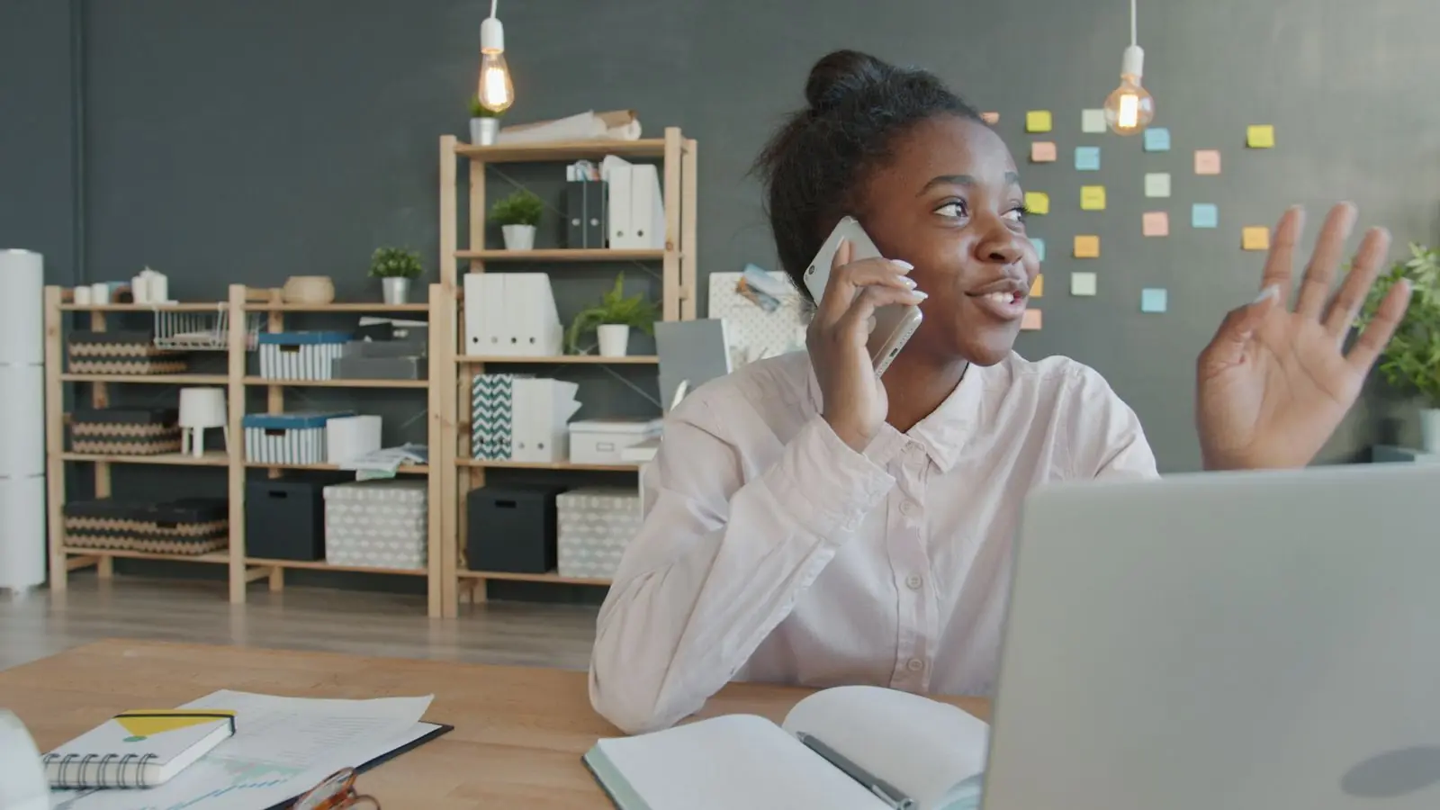 Une jeune femme souriante au téléphone dans son bureau, ordinateur et carnet devant elle